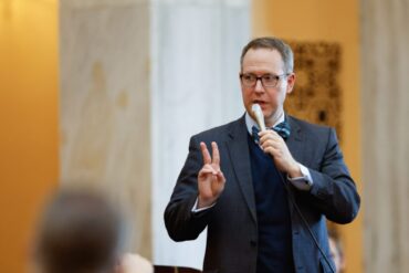 Photo of Ohio Sen Louis Blessing wearing a suit and talking into a microphone in a stately building